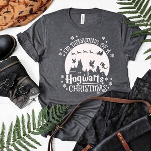 May include: A gray t-shirt with a white graphic design that says "I'm dreaming of a Hogwarts Christmas". The graphic features a moon, a castle, and flying creatures.