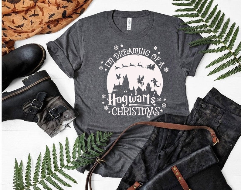 May include: A gray t-shirt with a white graphic design that says "I'm dreaming of a Hogwarts Christmas". The graphic features a moon, a castle, and flying creatures.