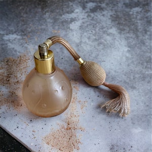 Puede incluir: Un frasco de perfume vintage de vidrio con un atomizador dorado y un borla marrón. El frasco tiene una superficie texturizada y está lleno de un líquido transparente.