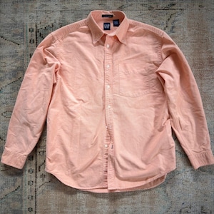 May include: A long-sleeve button-down shirt in a solid light peach color. The shirt features a pointed collar, a single chest pocket on the left side, and a front button placket. The interior neck label displays the brand name GAP in white text.
