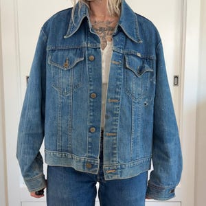 May include: A medium-wash blue denim jacket featuring a pointed collar, two button-flap chest pockets, and a front button closure. The jacket displays a worn texture with visible fading, light distressing, and a small hole on the left chest panel.