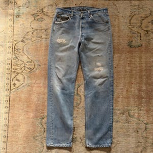 May include: Light blue denim jeans featuring a classic five-pocket design, a button fly closure, and a faded wash. The fabric displays intentional distressing with small frayed holes on the upper right thigh and the left knee area.