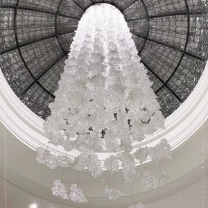 Puede incluir: Una gran lámpara de araña de cristal blanco cuelga de un techo circular de vidrio tintado. La lámpara de araña está compuesta por muchas piezas de cristal individuales con forma de hojas.