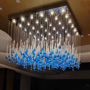 May include: A modern chandelier with a square frame and blue glass teardrop pendants. The chandelier is lit with white lights.