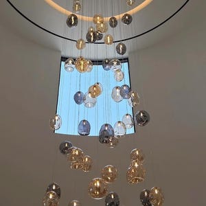 May include: A contemporary chandelier featuring numerous glass globe lights in amber, grey, and clear hues. The lights are suspended from the ceiling, forming a decorative and elegant lighting fixture. The fixture hangs in front of a window with a blue sky.