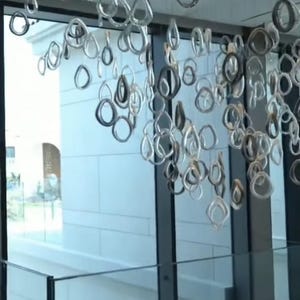 Puede incluir: Una lámpara de araña moderna compuesta por numerosos elementos de vidrio transparente de forma irregular suspendidos del techo. Las piezas de vidrio varían en tamaño y forma, creando una exhibición dinámica y artística. La lámpara de araña cuelga frente a una ventana grande.