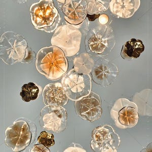 May include: A ceiling light fixture featuring numerous suspended glass elements shaped like flowers. The pieces vary in color, ranging from clear and translucent white to warm amber and deep bronze tones, creating a layered, floating floral arrangement.