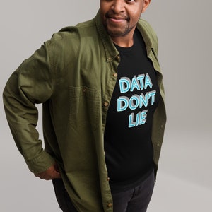 Può includere: Un uomo indossa una maglietta nera con la scritta "DATA DON'T LIE" in lettere blu e bianche. Indossa una camicia verde abbottonata sopra la maglietta e jeans neri. Indossa scarpe da ginnastica verdi.