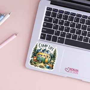 May include: A sticker featuring a vintage camper van parked in a forest setting with mountains in the background. The sticker says "Camp Life" and includes a campfire, a lantern, and a chair. The sticker is on a laptop.