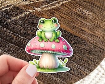 Könnte beinhalten: Ein gestanzter Aufkleber mit einem kleinen grünen Frosch mit großen Augen, der auf einem rosa Pilz mit weißen Punkten sitzt. Der Pilz hat einen hellen Stiel und grünes Gras an der Basis. Der Text KENJIA DESIGN ist oben rechts zu sehen.