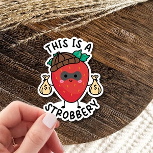 May include: A sticker with a cartoon strawberry wearing a brown hat and a black mask, holding two bags of money. The text "This is a strobbery" is written below the strawberry.