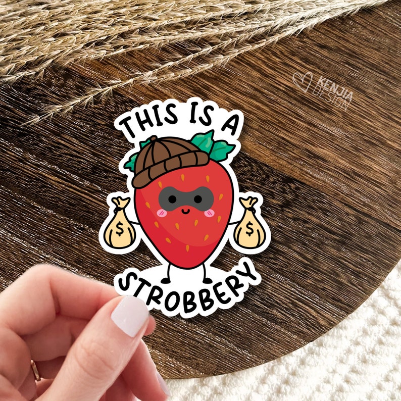May include: A sticker with a cartoon strawberry wearing a brown hat and a black mask, holding two bags of money. The text "This is a strobbery" is written below the strawberry.