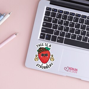 May include: A red strawberry with a black eye mask and a brown hat is holding two bags of money. The text "This is a Strobbery" is written below the strawberry.