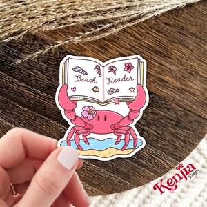 Op de afbeelding: Een roze krabsticker met een boek met de tekst "Beach Reader". Het boek heeft illustraties van schelpen, zeepaardjes, bloemen en vissen. De krab staat op een blauw en geel strand.