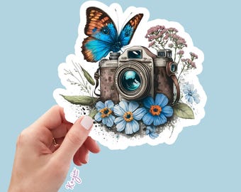 May include: A die-cut sticker featuring a vintage-style camera in brown and silver tones, adorned with a large blue and orange butterfly, light blue flowers with orange centres, and small pink floral sprigs. The text Kenjia design is visible.
