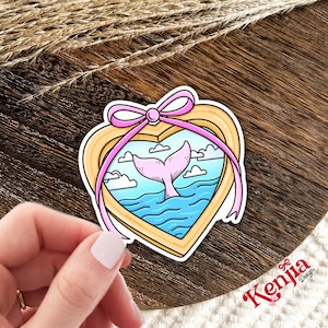 May include: A heart-shaped sticker featuring a pink whale tail in blue ocean waters under a cloudy sky. The sticker is framed with a gold border and a pink ribbon. The sticker has the text "Kenjia Design".