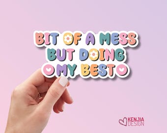 May include: A die-cut sticker featuring the text BIT OF A MESS BUT DOING MY BESTO in a retro, multi-colored font. The letters are colored in shades of pink, yellow, blue, and purple, with small white flower icons replacing the letter O.
