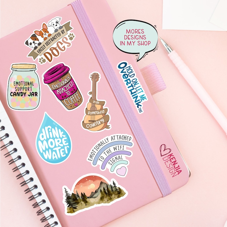 May include: A collection of colorful stickers featuring various designs, including a jar with candy, a coffee cup, a guitar, a water drop, a wifi signal, a mountain scene, and a heart with the text "Kenjia Design". The stickers are on a pink notebook with a spiral binding.