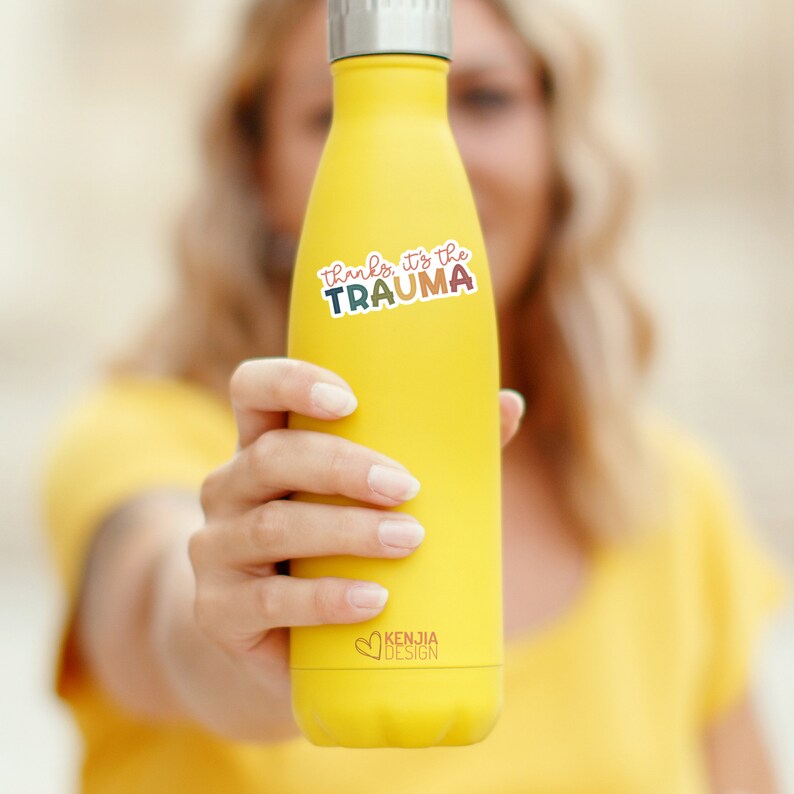 May include: A yellow reusable water bottle with a sticker that says "Thanks, it's the trauma" in rainbow colors. The bottle has a silver lid and a small heart logo that says "Kenjia Design".