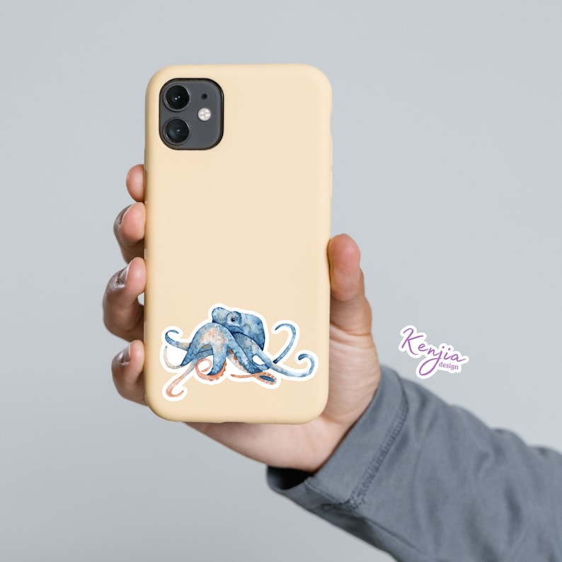 May include: A light yellow phone case with a blue and pink watercolor octopus illustration. The octopus has eight tentacles and is curled up in a relaxed position. The phone case has a camera cutout and a white border around the octopus illustration. The text "Kenjia Designs" is in purple text on the bottom right corner of the phone case.