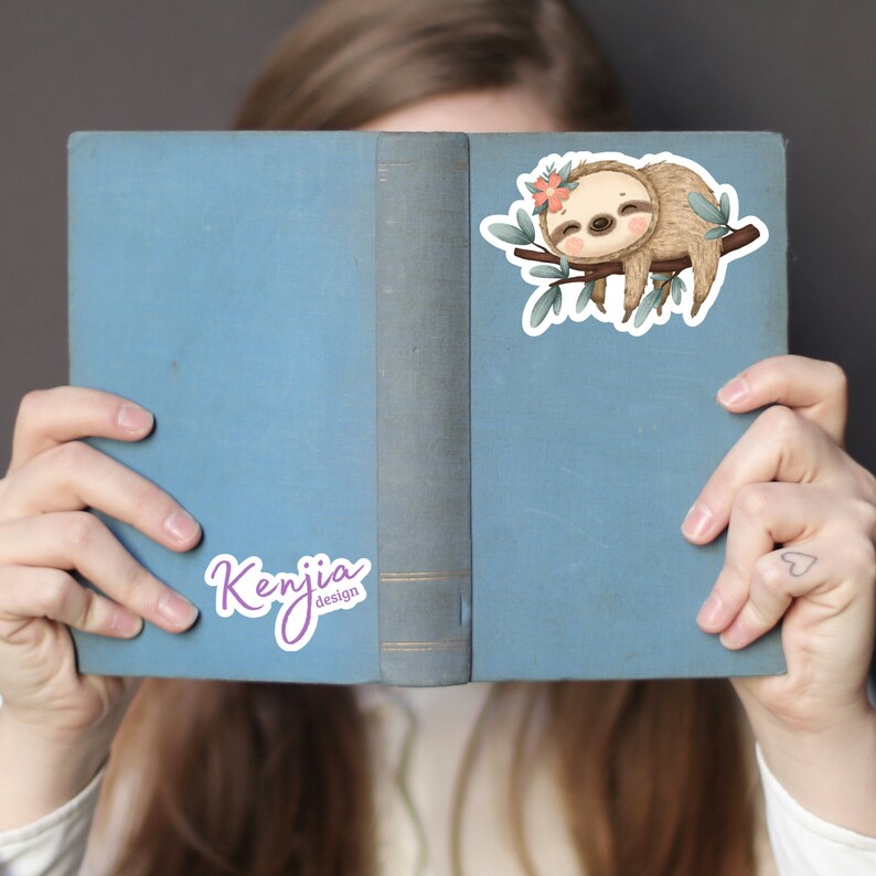 May include: A blue book with a sloth sticker on the cover. The sticker features a sloth with a flower crown, sleeping on a branch. The book also has a sticker with the text "Kenjia design" on the cover.