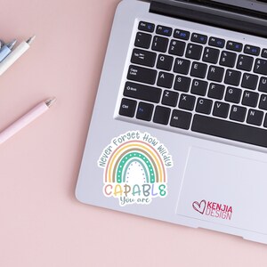 Op de afbeelding: Een laptop met een sticker met een regenboogontwerp en de woorden "Never Forget How Wildly CAPABLE You are." De sticker heeft een pastel kleurenschema. Een roze pen en twee andere pennen liggen op een roze oppervlak.
