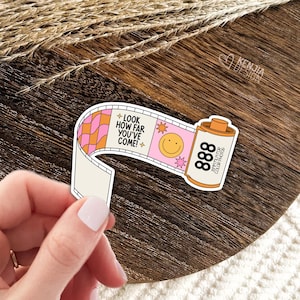 May include: A pink and orange film strip sticker with a smiley face and the text "LOOK HOW FAR YOU'VE COME!" and "35MM FILM FOR COLOR PHOTOS" on a brown wooden surface.