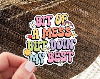 May include: A die-cut sticker featuring the text "BIT OF A MESS BUT DOIN' MY BEST" in colorful, bubbly, retro-style typography. The design includes small graphic elements like a rainbow, a smiley face, white flowers, and a winged heart.