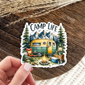 May include: A sticker featuring a vintage camper van parked in a forest setting with a campfire, a folding chair, and a lantern. The text "Camp Life" is written in a retro font above the camper.