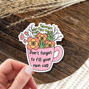 May include: A die-cut sticker featuring a pink mug with horizontal stripes, filled with a bouquet of orange, yellow, and green flowers. The text "Don't forget to fill your own cup" is printed in black on the front of the mug in a simple font.