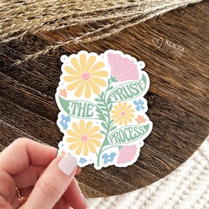 May include: A sticker featuring a floral design and the text "Trust the Process". The flowers are yellow, pink and blue. The text is green.