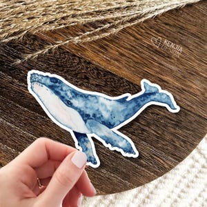 May include: A die-cut sticker featuring a watercolor illustration of a blue humpback whale with a white underbelly. The whale is depicted in profile with mottled blue tones. The text KENJIA DESIGN is visible in the upper right corner.
