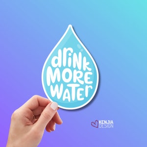 Könnte beinhalten: Ein hellblauer Aufkleber in Tropfenform mit dem weißen Schriftzug "drink MORE WATER". Der Aufkleber wird von einer Hand vor einem blau-violetten Farbverlaufshintergrund gehalten. Das Kenjia Design Logo befindet sich unten rechts.