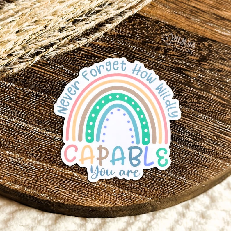 Op de afbeelding: Een kleurrijke sticker met een regenboogontwerp en de tekst "Never Forget How Wildly CAPABLE You are." De sticker heeft pastelkleuren, waaronder roze, oranje, groen en blauw. De sticker ligt op een houten oppervlak.