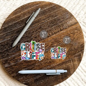 May include: Two vinyl stickers with the text "Cruise Life" in colorful letters with a beachy design. The stickers are shown on a wooden surface with a pen next to them. The larger sticker is 3 inches and the smaller sticker is 2 inches.