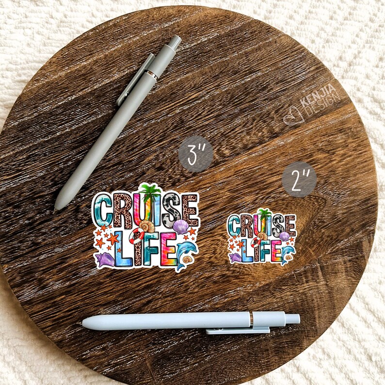 May include: Two vinyl stickers with the text "Cruise Life" in colorful letters with a beachy design. The stickers are shown on a wooden surface with a pen next to them. The larger sticker is 3 inches and the smaller sticker is 2 inches.