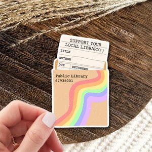Puede incluir: Una pegatina con un diseño de arcoíris y el texto "¡Apoya tu biblioteca local!" y "Biblioteca Pública 67938001". La pegatina tiene forma de bolsillo de tarjeta de biblioteca con una tarjeta dentro.