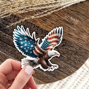 May include: A die-cut sticker featuring a bald eagle in flight with wings spread wide. The eagle's feathers are patterned with the red, white, and blue colors of the American flag, including stars on the left wing. The text KENJIA DESIGN is visible.