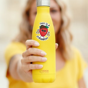 May include: Yellow reusable water bottle with a sticker of a cartoon strawberry wearing a hat and sunglasses and holding money bags. The sticker says "This is a Strobbbery".