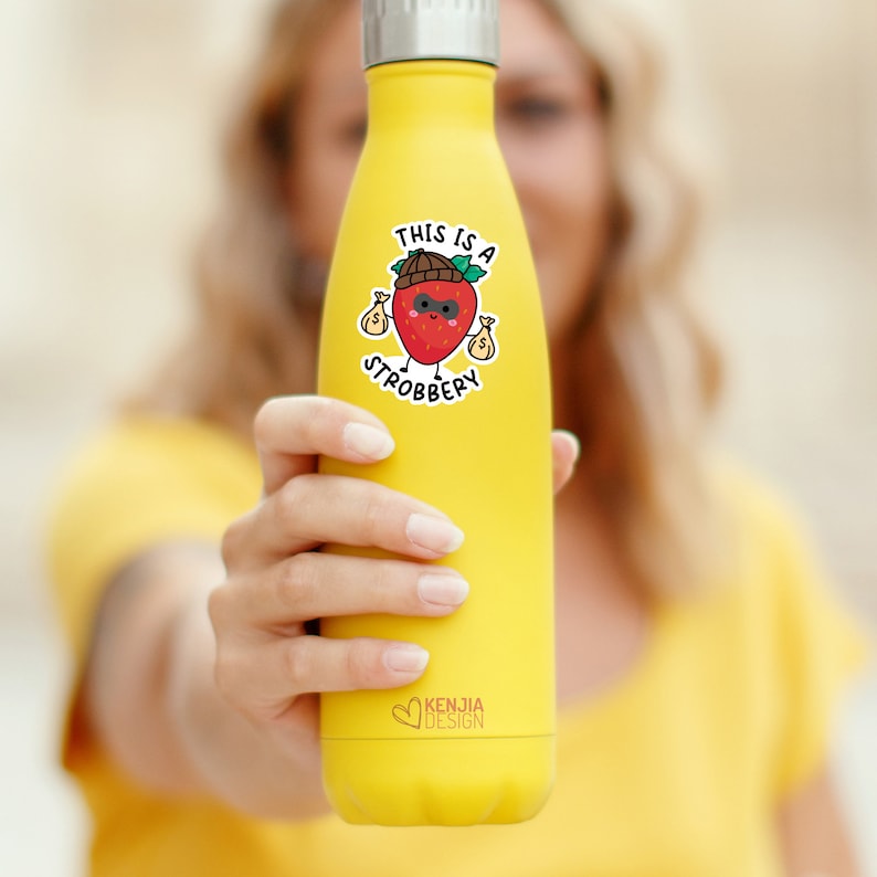 May include: Yellow reusable water bottle with a sticker of a cartoon strawberry wearing a hat and sunglasses and holding money bags. The sticker says "This is a Strobbbery".