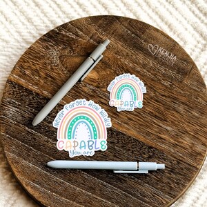 Op de afbeelding: Twee lichtgrijze pennen en twee stickers met een regenboogontwerp en de tekst "Never Forget How Wildly CAPABLE You are" worden weergegeven op een houten oppervlak. De stickers hebben pastelkleuren.
