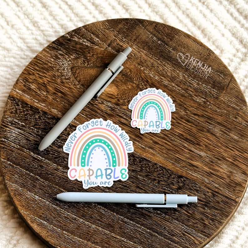 Op de afbeelding: Twee lichtgrijze pennen en twee stickers met een regenboogontwerp en de tekst "Never Forget How Wildly CAPABLE You are" worden weergegeven op een houten oppervlak. De stickers hebben pastelkleuren.