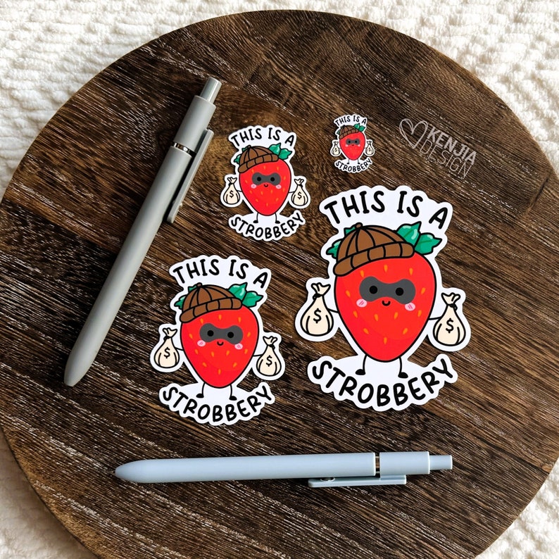 May include: Three stickers featuring a cartoon strawberry wearing a robber hat and holding bags of money. The text on the stickers reads "This is a Strobbbery".