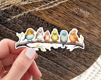 May include: A die-cut sticker featuring six colourful cartoon birds perched on a brown tree branch. The birds are orange, light blue, yellow, pink, grey, and a mix of orange and blue. The text KENJIA DESIGN is printed in the upper right corner.