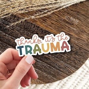 May include: A sticker with the text "thanks, it's the trauma" in rainbow colors with a white outline. The sticker is on a wooden surface.