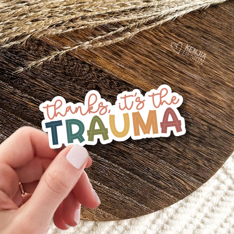 May include: A sticker with the text "thanks, it's the trauma" in rainbow colors with a white outline. The sticker is on a wooden surface.