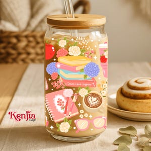 Può includere: Un bicchiere di vetro trasparente con coperchio e cannuccia in bambù, pieno di una bevanda. Il bicchiere è decorato con un design colorato con libri, fiori, una tazza di caffè e la scritta "Teach Love Inspire". Il logo Kenjia Design è visibile.