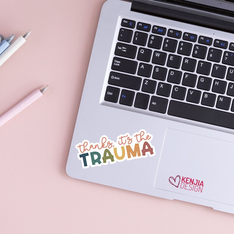 May include: A laptop computer with a sticker that says "thanks, it's the trauma" in colorful letters. The sticker is on the trackpad of the laptop. The laptop is on a pink surface.
