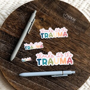 May include: Four colorful stickers with the text "thanks, it's the trauma" in different color combinations. The stickers are on a wooden surface with two pens.