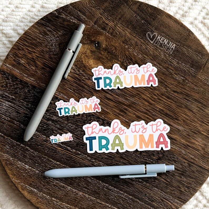 May include: Four colorful stickers with the text "thanks, it's the trauma" in different color combinations. The stickers are on a wooden surface with two pens.